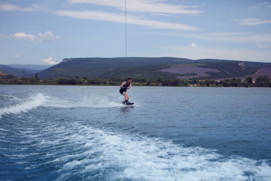 Young sportsman surfing across lake. Surfer wearing wet swimsuit training in wake park, wakeboarding on river, pulled by motor boat, clinging to cable. Wakesurfing, waterskiing, sports and recreation